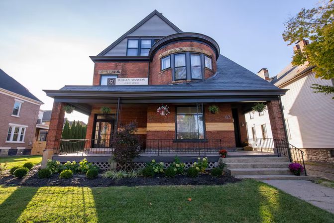 Historic red-brick Victorian house with a rounded turret, wide covered porch with hanging flower baskets, stone steps and a manicured front lawn under a clear blue sky.