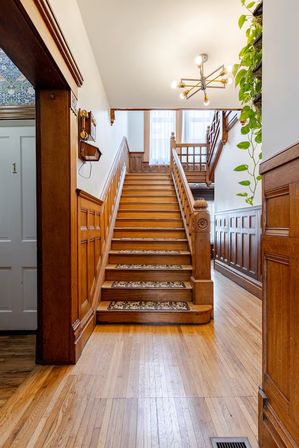 Sunlit wood-paneled entry with a wide oak staircase, patterned stair runners, modern chandelier and trailing pothos in a historic townhouse