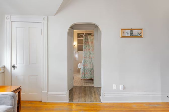 Bright apartment interior with warm hardwood floors, white paneled door and arched doorway leading to a bathroom with a pastel floral shower curtain and a small framed print on a pale gray wall.