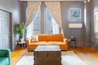 Sunlit urban apartment living room with a mustard-orange tufted sofa, patterned curtains framing tall windows, vintage trunk coffee table on a light rug, gray walls, hardwood floors, potted plant and wall-mounted air conditioner.