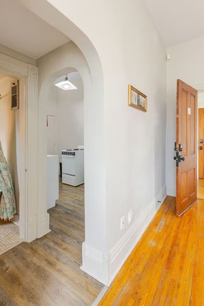 Bright apartment entryway with an open wooden front door, arched doorway to a compact kitchen with a white stove, mixed hardwood and vinyl plank floors, and pale gray walls.