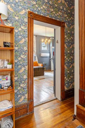 Peek through an ornate wooden doorway framed by blue floral-and-bird wallpaper into a cozy vintage living room with hardwood floors, a brass chandelier, orange armchair, and trunk-style coffee table.