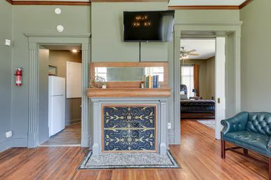 Residential living room with decorative wooden mantel and ornate metal fireplace cover, wall-mounted flat-screen TV, sage green walls, tufted leather armchair, hardwood floors, and doorways to a kitchen and adjoining sitting room.