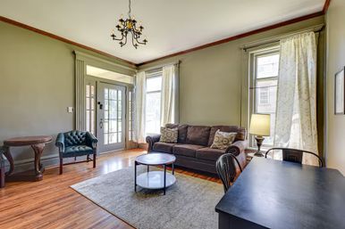Cozy sunlit living room with hardwood floors and classic trim, featuring a brown upholstered sofa with patterned pillows, tufted leather armchair, round two-tier coffee table on a gray area rug, chandelier, tall windows with sheer curtains, and a front entry door with sidelights.