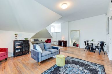 Bright loft-style open-concept living space with sloped ceiling and hardwood floors, blue velvet loveseat, patterned rug and green ottoman, red accent chair, small dining nook with black metal chairs, full-length mirror and dormer window