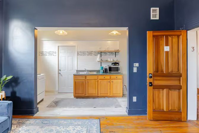 Cozy sunlit studio interior with navy blue walls and hardwood floors, open kitchenette featuring wooden cabinets, sink and microwave, wooden entry door, area rug and small potted plant