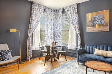 Sunlit cozy urban living room with a bay window framed by blue floral curtains, round metal dining table with four chairs, navy tufted velvet sofa, patterned rug and hardwood floors.