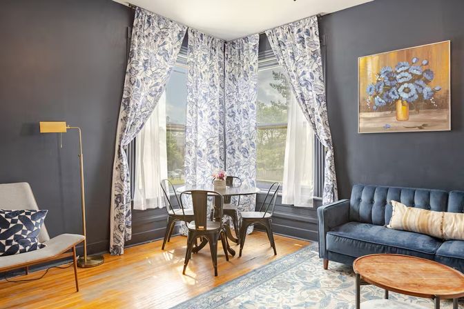 Sunlit cozy urban living room with a bay window framed by blue floral curtains, round metal dining table with four chairs, navy tufted velvet sofa, patterned rug and hardwood floors.