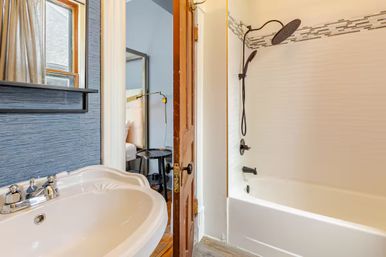 Cozy small home bathroom with white sink and tub, dark bronze rain showerhead, blue textured wall and open wooden door revealing a bedroom with bedside table and lamp.