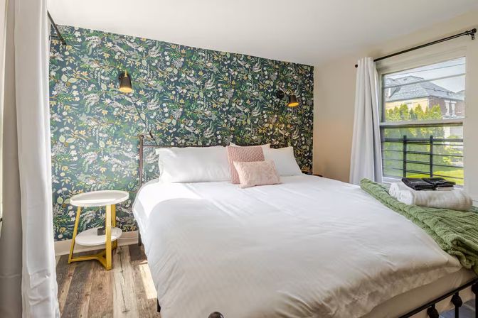 Cozy sunlit bedroom with a king bed in crisp white linens, green botanical accent wallpaper, brass wall lamps, round gold-and-white bedside table, hardwood floors, and a window view of the neighborhood.