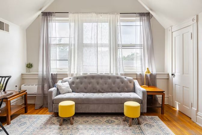 Bright upstairs living room with a gray tufted sofa, two yellow ottomans, sheer window curtains, wooden side tables and hardwood floors.