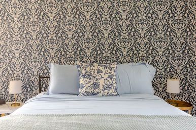 Cozy chic bedroom interior with navy-and-cream damask wallpaper featuring stag motifs, bed dressed in pale blue linens with a blue floral accent pillow and brass bedside lamps on round tables.