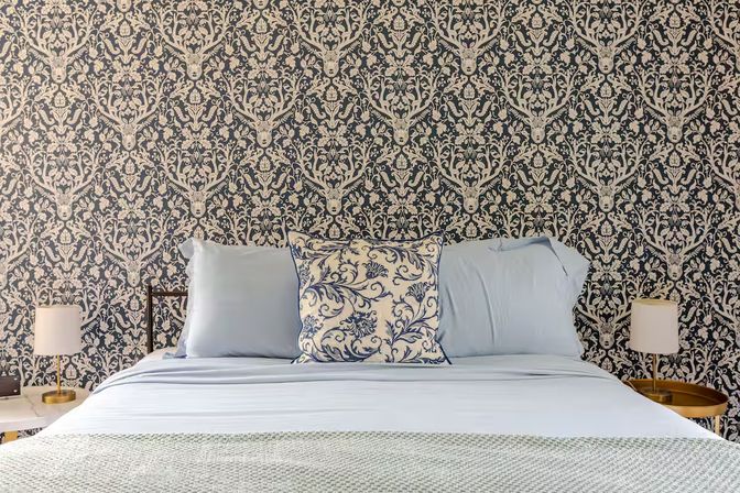 Cozy chic bedroom interior with navy-and-cream damask wallpaper featuring stag motifs, bed dressed in pale blue linens with a blue floral accent pillow and brass bedside lamps on round tables.