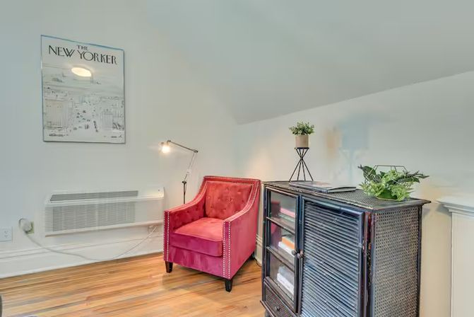 Cozy attic reading nook in a home with a red tufted armchair, adjustable floor lamp, framed magazine cover on the wall, dark woven cabinet topped with potted plants, and warm hardwood floors.