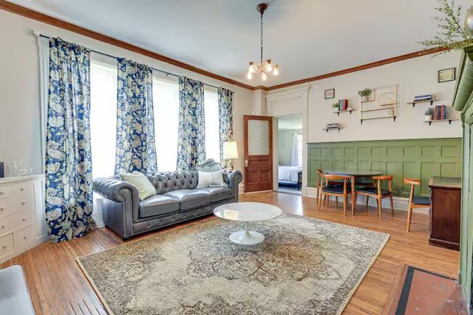 Cozy sunlit living room with tall windows and blue floral curtains, tufted gray leather sofa, white round coffee table, patterned area rug, green paneled wainscoting and hardwood floors.