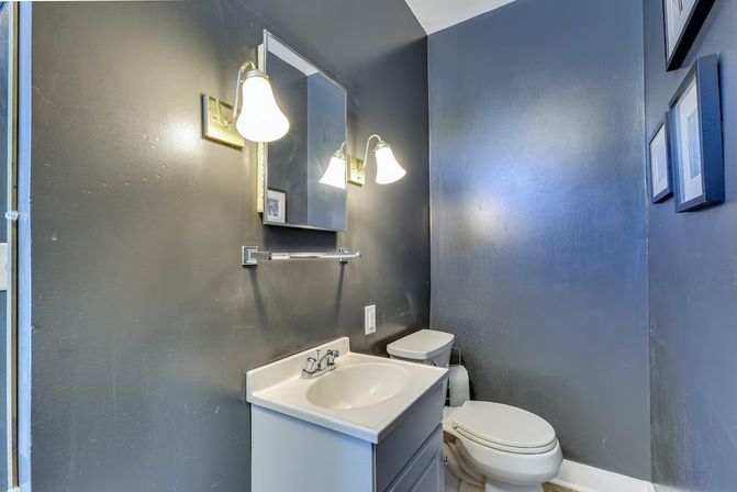 Compact residential powder room with dark gray walls, white vanity sink, wall-mounted mirror, two lit brass sconces, and a white toilet.