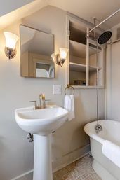 Cozy small bathroom with a white pedestal sink, clawfoot tub and overhead rain shower, mirrored cabinet, open shelves and glowing wall sconces