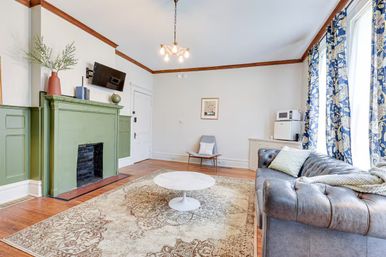 Sunny Victorian-style living room with green painted fireplace, tufted gray leather sofa, round white coffee table on an ornate area rug, hardwood floors, blue floral curtains and wall-mounted TV.