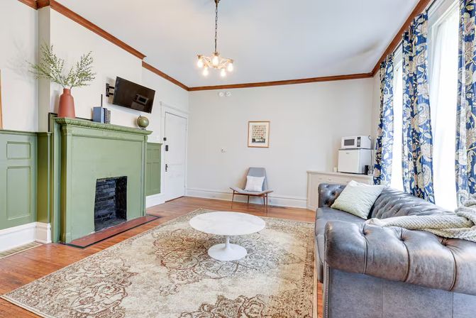 Sunny Victorian-style living room with green painted fireplace, tufted gray leather sofa, round white coffee table on an ornate area rug, hardwood floors, blue floral curtains and wall-mounted TV.