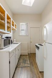 Cozy narrow galley apartment kitchen with white cabinets, open yellow-backed shelves, microwave, sink, compact electric stove, refrigerator, wood-look floor and small window.