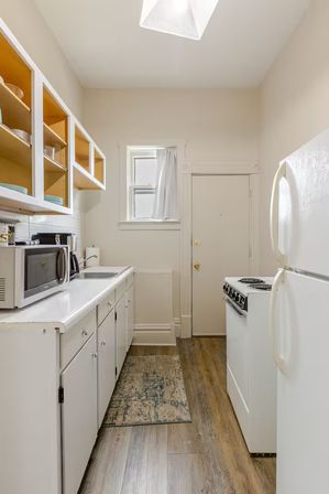 Cozy narrow galley apartment kitchen with white cabinets, open yellow-backed shelves, microwave, sink, compact electric stove, refrigerator, wood-look floor and small window.