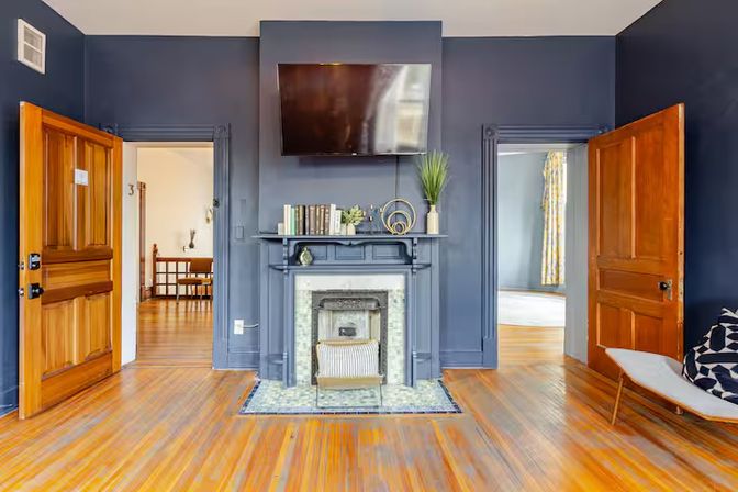 Cozy navy-blue living room with hardwood floors, decorative fireplace and mounted flat-screen TV, mantle styled with books and a plant, and open wooden doors revealing adjacent rooms.