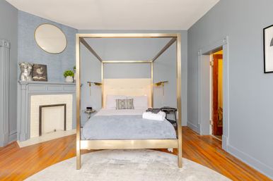 Stylish bedroom interior with a gold four-poster canopy bed, light gray-blue walls, decorative fireplace topped by a round mirror, hardwood floors, brass bedside lamps, and folded towels on the bed.