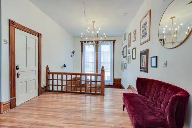 Sunlit upstairs landing in a renovated home with polished hardwood floors, decorative wood railing, brass chandelier, gallery wall and a cozy burgundy velvet sofa.