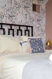 Cozy modern bedroom interior with black metal headboard, pale pink floral wallpaper, cream bedding, patterned throw pillows and brass bedside lamp