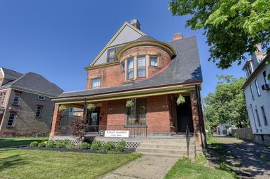 Historic red-brick Victorian house with rounded bay window and covered front porch, hanging fern planters, front steps and neat lawn under a clear blue sky in a residential neighborhood.