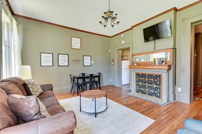 Sunlit cozy living room with brown leather sofa, round coffee table, hardwood floors, ornate decorative fireplace with iron grille, wall-mounted TV, small black dining table and chairs, chandelier, and large windows