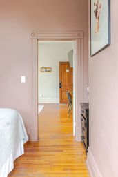 Peek-through view from a blush-pink bedroom into an adjoining room with polished hardwood floors, decorative door trim, wooden door, wall-mounted TV, white bedspread and black dresser.