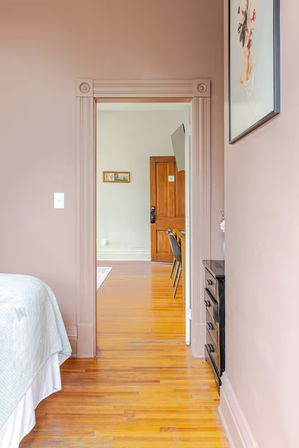 Peek-through view from a blush-pink bedroom into an adjoining room with polished hardwood floors, decorative door trim, wooden door, wall-mounted TV, white bedspread and black dresser.