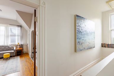 Sunlit upstairs landing opening to a cozy living area with hardwood floors, gray tufted sofa, yellow ottoman, wooden side table with lamp, abstract wall art and books on the windowsill.