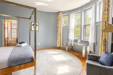 Sunny bedroom with brass four-poster bed, gray walls, curved bay windows with yellow floral curtains and sheer panels, round light rug on hardwood floors, glass console table with flowers and a gray velvet armchair.