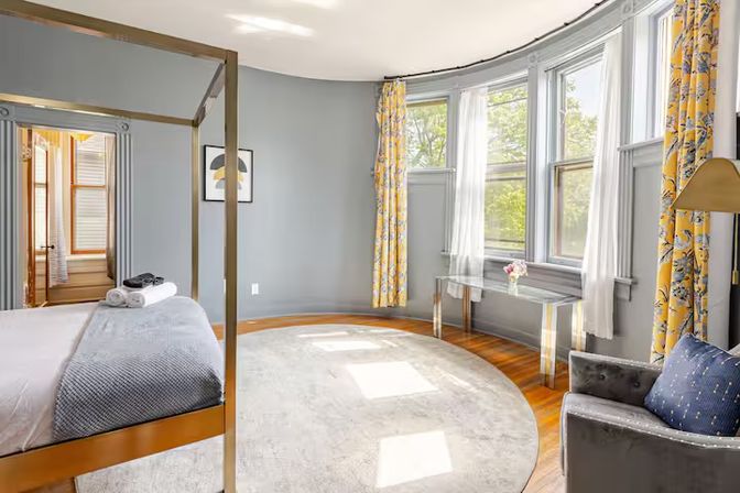 Sunny bedroom with brass four-poster bed, gray walls, curved bay windows with yellow floral curtains and sheer panels, round light rug on hardwood floors, glass console table with flowers and a gray velvet armchair.