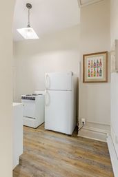 Cozy small apartment kitchen with white top-freezer refrigerator and compact electric stove, wood-look vinyl plank floor, beige walls, pendant light and framed colorful bottle art.