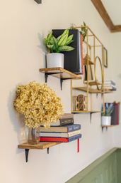 Floating wooden shelves on a cream living room wall with potted green plant, gold geometric shelving, stacked books and a glass vase of dried hydrangea — cozy home decor