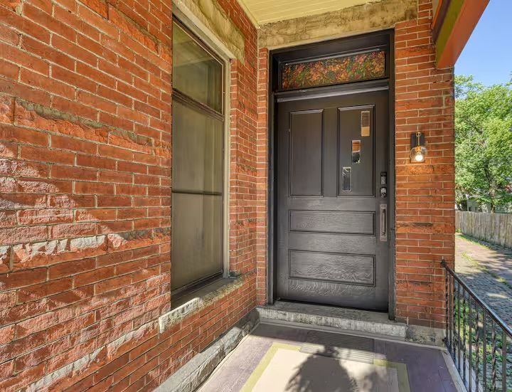 Cozy red-brick porch with a matte black wooden front door, decorative stained-glass transom above, a single wall lantern, stone step and iron railing leading to a tree-lined sidewalk.