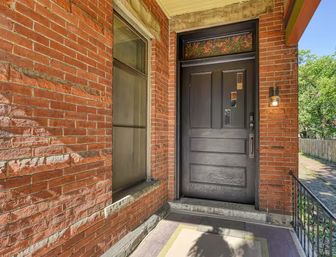 Cozy red-brick porch with a matte black wooden front door, decorative stained-glass transom above, a single wall lantern, stone step and iron railing leading to a tree-lined sidewalk.