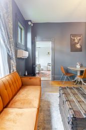Cozy chic living room with orange velvet sofa, rustic wooden trunk coffee table, gray walls with a bull portrait, round dining table with orange chairs, floral curtains and a doorway leading to a bedroom with a patterned rug.