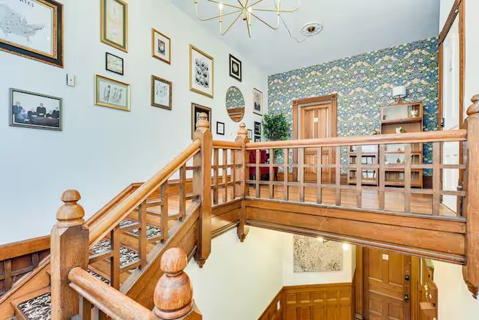 Sunlit Victorian-style interior with a carved wooden staircase and upstairs landing, spindle banister, gallery wall of framed art, floral wallpaper, chandelier and wooden doors