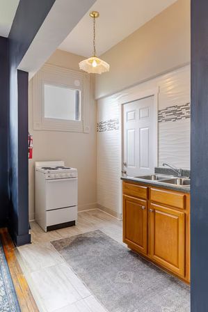 Cozy narrow galley kitchen with compact white stove, oak cabinets, double stainless sink, mosaic tile backsplash, pendant light and gray runner rug.