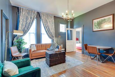 Bright, stylish living room with blue-gray walls and hardwood floors, tufted tan sofa and emerald armchair, patterned curtains over large windows, vintage trunk coffee table and small dining nook