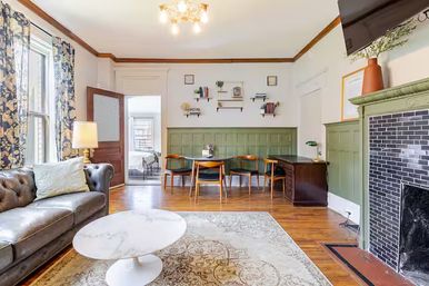 Sunny period-style living room in a townhouse with tufted leather sofa, round marble coffee table, patterned curtains, green wainscoting, black-tiled fireplace, hardwood floors, and a small dining nook.