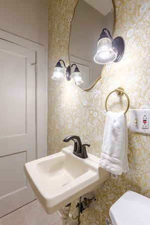Cozy vintage-inspired powder room with yellow floral wallpaper, scalloped oval mirror, twin glass wall sconces, cream wall-mounted sink with matte black faucet, brass towel ring holding a white hand towel, and tiled floor.