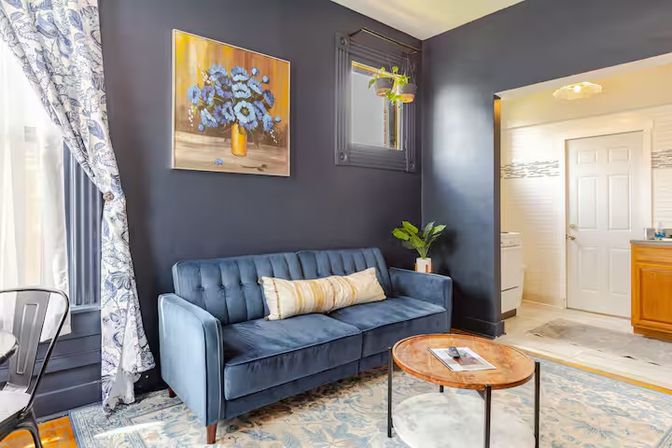Cozy modern living room in a small apartment with a navy accent wall, plush velvet blue sofa, painting of blue flowers in a yellow vase, round wooden coffee table on a patterned rug, potted plant and floral curtains near a doorway to a bathroom.