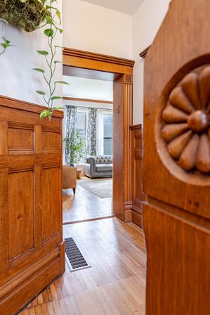 Sunlit craftsman-style hallway with warm oak wainscoting and carved door detail opening to a living room with a tufted gray sofa, floral curtains, hardwood floors, and indoor plants.