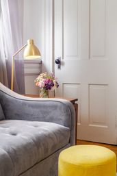 Cozy living room corner with gray velvet sofa, bright yellow round ottoman, brass floor lamp and glass vase of mixed flowers on a wooden side table by a white paneled door.