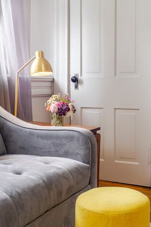 Cozy living room corner with gray velvet sofa, bright yellow round ottoman, brass floor lamp and glass vase of mixed flowers on a wooden side table by a white paneled door.